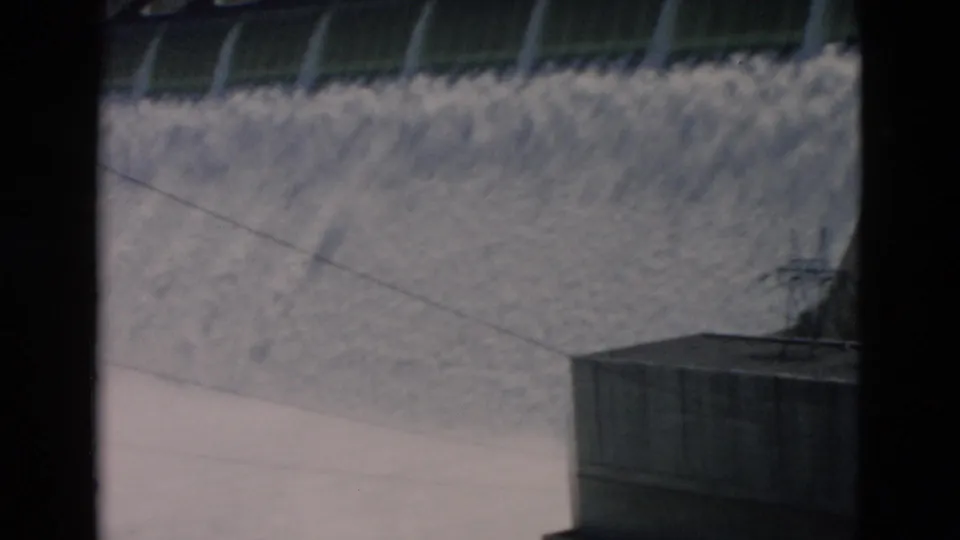 1958 8mm Montana footage shows water pouring over a concrete dam, with a structure and film-frame borders visible.