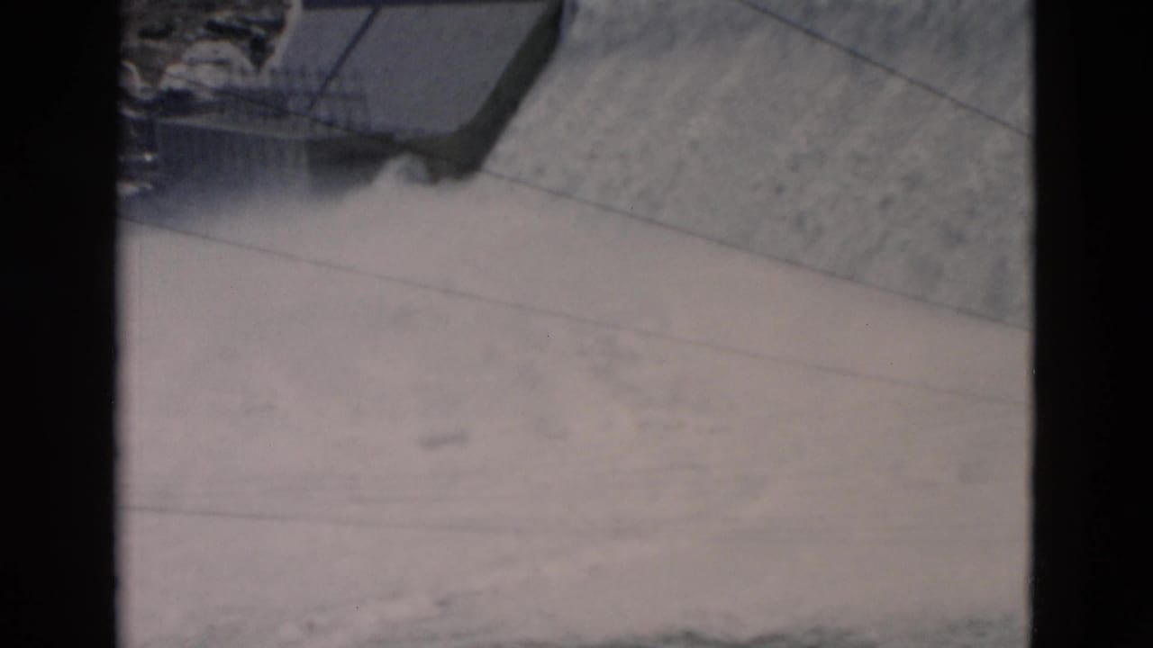 1958 Montana dam footage shows a concrete spillway edge, misting water, railings, and rocky ground above the structure.