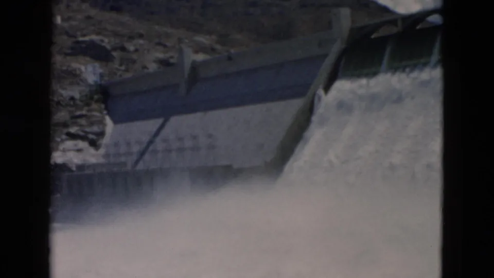 1958 8mm Montana frame shows water rushing over a concrete dam wall, with railings, rocks, and soft film grain.