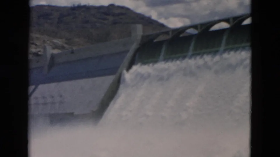 1958 Montana home-movie footage shows white water pouring over a concrete dam beneath rocky hills and a cloudy sky.