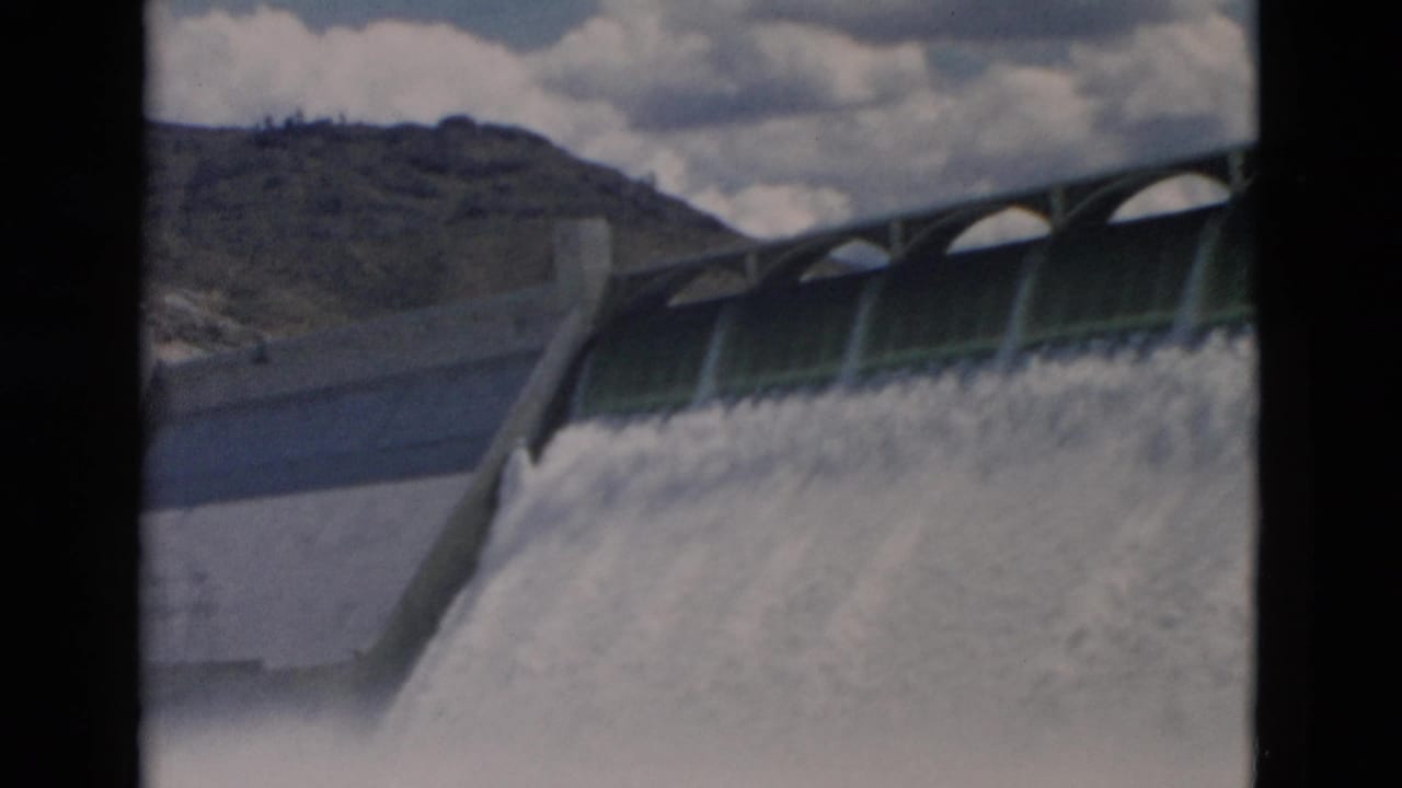 Concrete dam spillway releasing a broad cascade of white water below a cloudy sky.