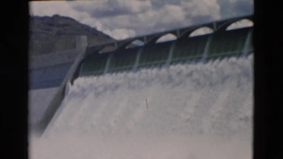 1958 Montana 8mm view of a dam spillway releasing a dense sheet of white water, with hills and clouds beyond.