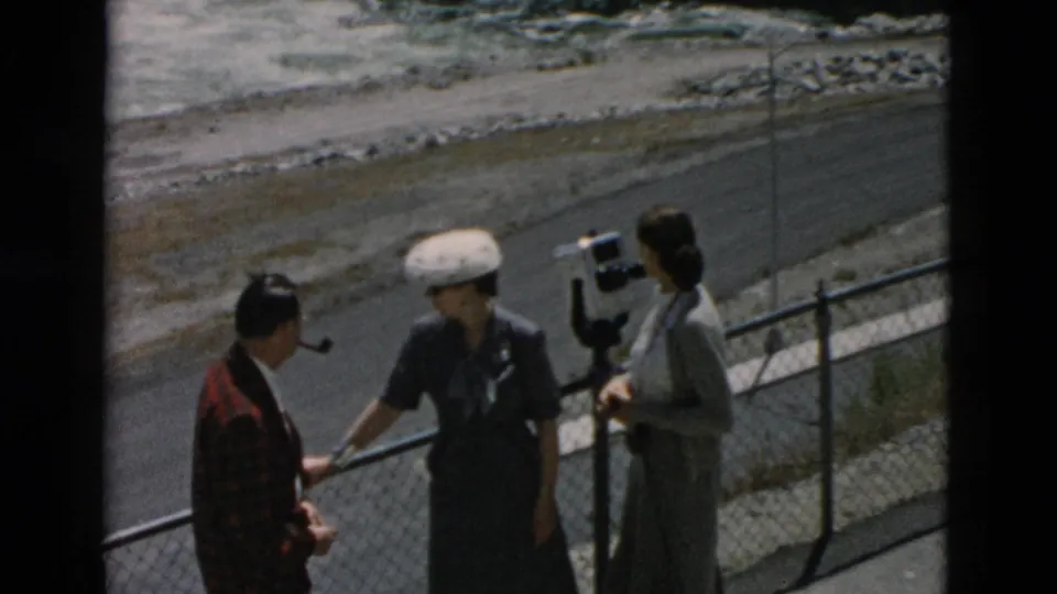 Three women in 1950s attire stand at a Montana overlook fence, one using a coin-operated viewer near the dam.