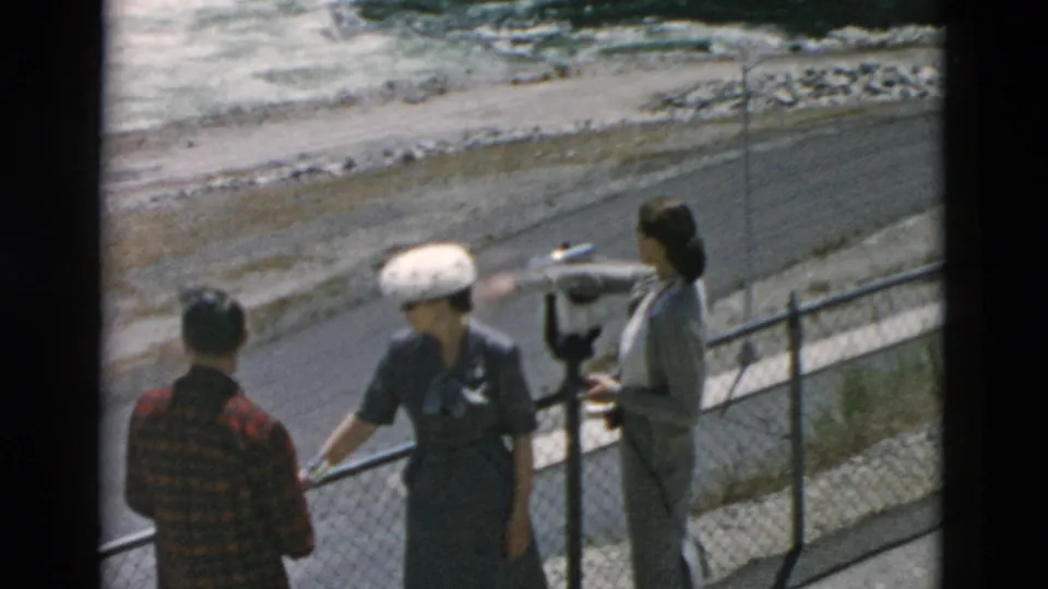 Three adults at a Montana overlook in 1958 stand near a coin-operated viewer as water rushes beyond the fence.