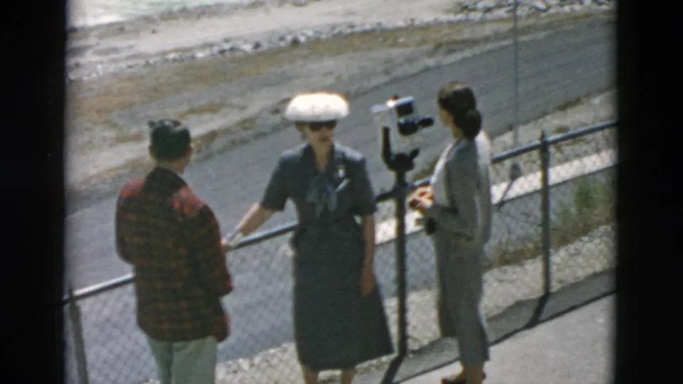 Three visitors at a 1958 Montana overlook stand near the chain-link fence, viewer, and rushing water.