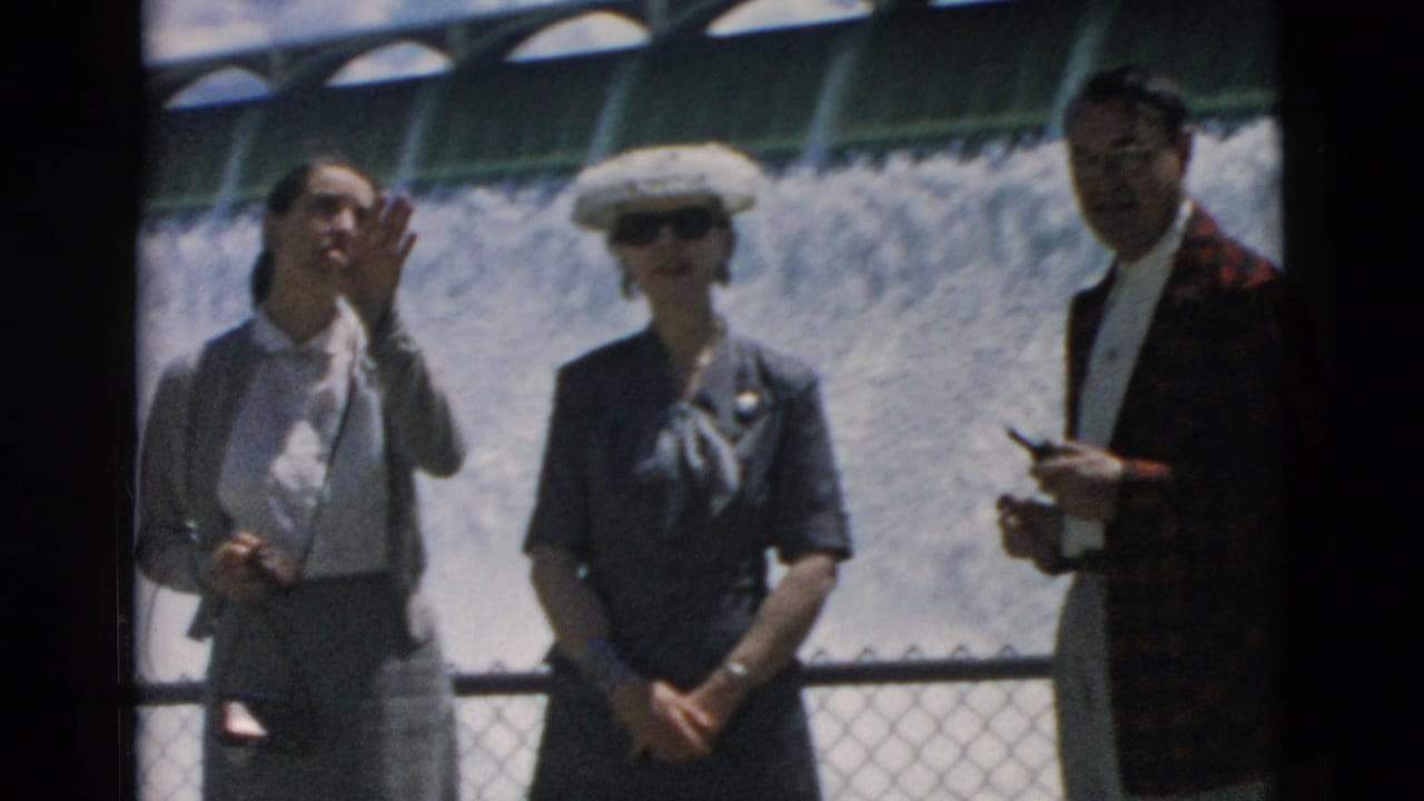 Three adult tourists stand in front of a dam water cascade, with one person waving toward the camera.