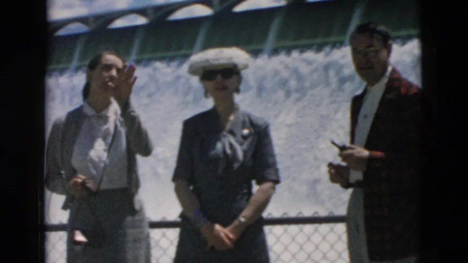 1958 Montana home-movie frame showing three adult tourists by a railing with a dam's water cascade and arched spillway behind them.