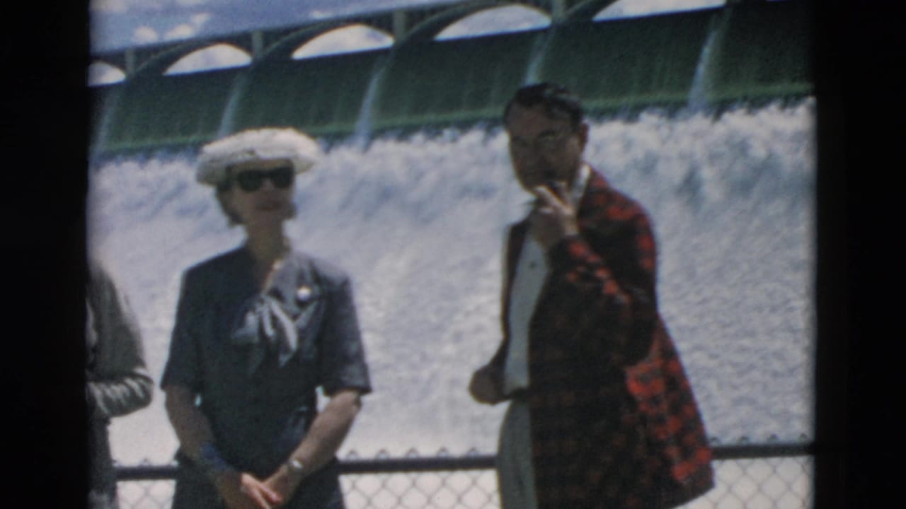 Two adult tourists stand before the dam, one wearing a white hat and another holding a cigarette.