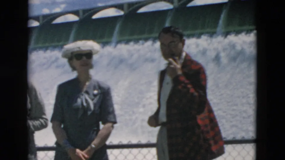 1958 Montana home-movie frame with two adults near a dam railing, bright spillway water filling the background.