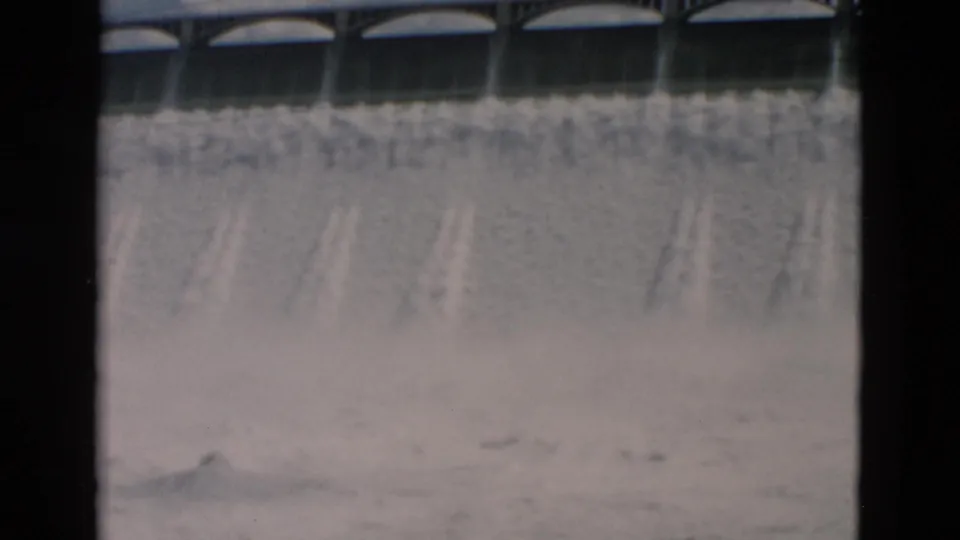1958 Montana home-movie frame emphasizing the dam's arched spillway and broad curtain of rushing water.
