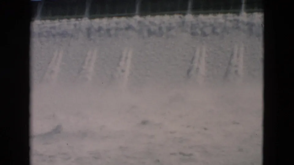 1958 Montana home-movie frame showing water cascading over a concrete dam spillway with mist below.
