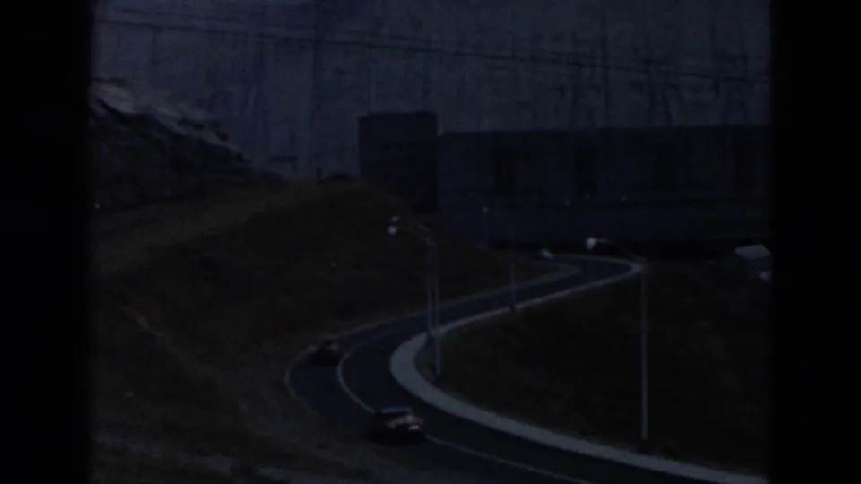 Dim 1958 Montana roadside view with a curved road, a car, utility poles, and a large dam structure in the background.