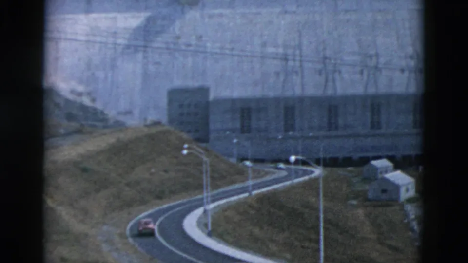 1958 Montana home-movie frame of a car on a winding road near a concrete dam, with poles, hillside, and power lines visible.