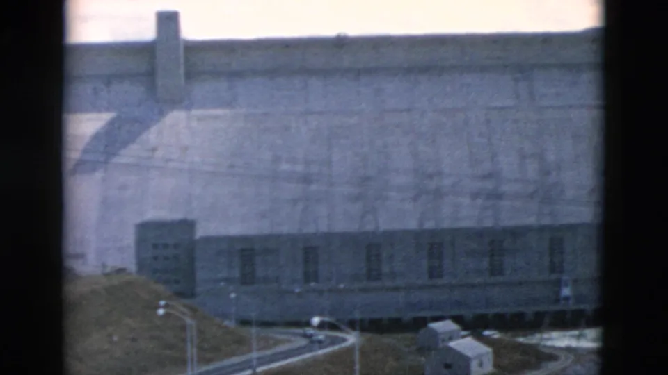 1958 Montana view of a concrete dam complex with road curves, small buildings, utility poles, and a high dam wall.