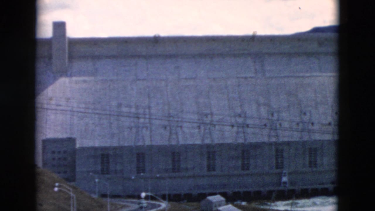 Distant concrete dam face with utility wires, road, hills, and cloudy sky.