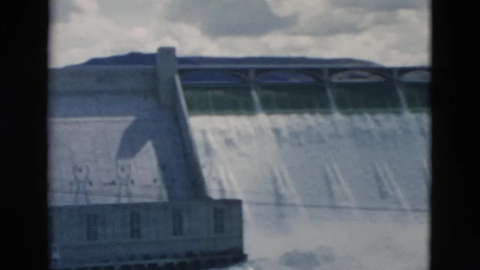 1958 Montana 8mm frame showing a wide spillway release, concrete dam structures, mist, utility wires, and cloudy sky.