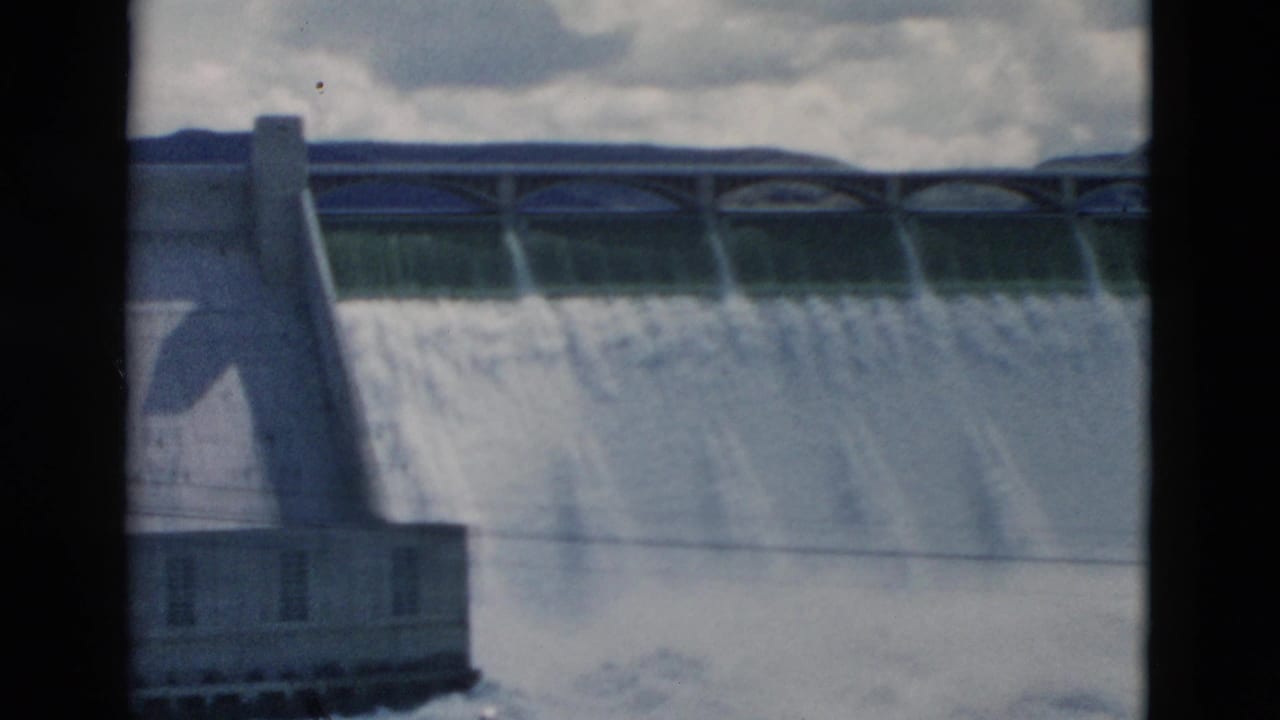 Dam spillway releasing heavy water beneath a cloudy sky, with an arched bridge across the top.