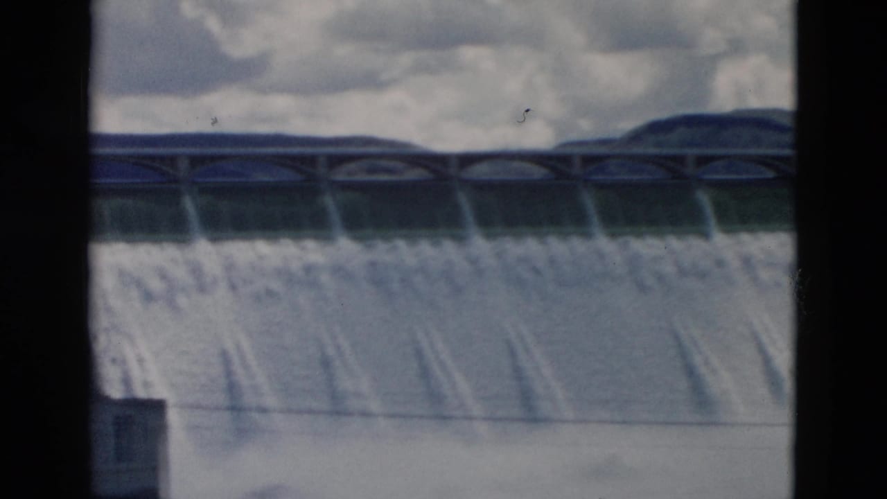 Wide view of water cascading over a dam with clouds and hills in the background.