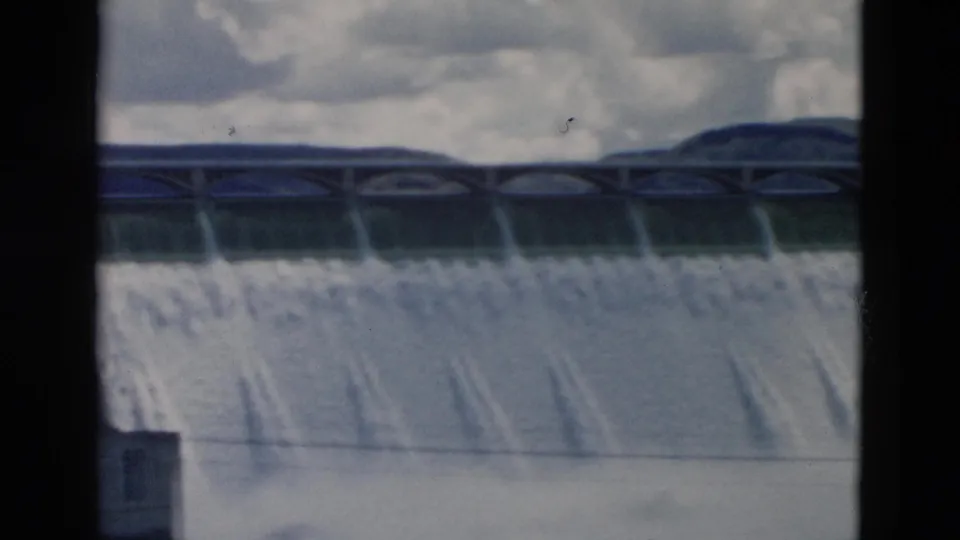 1958 Montana home-movie frame of water cascading over a broad dam, with an arched top structure, distant hills, cloudy sky, and film-frame edges.