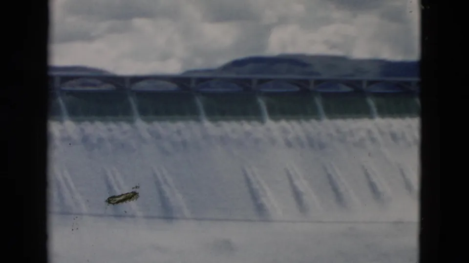 Soft 1958 Montana 8mm view of a dam spillway, arched bridge or roadway, heavy flowing water, cloudy sky, and visible film wear near the lower edge.