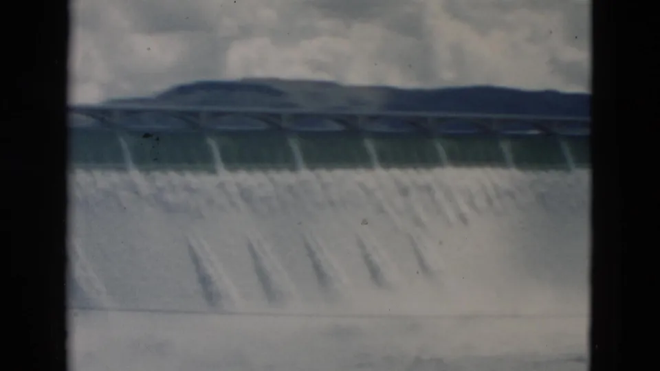 1958 Montana 8mm frame showing heavy water rushing over a dam, an arched bridge-like structure at the crest, distant hills, cloudy sky, and dark frame borders.