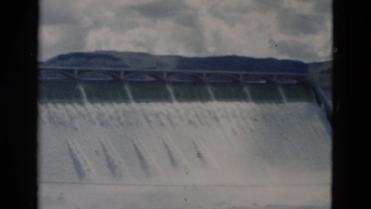 Dam spillway with water flowing under a cloudy sky.