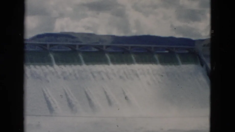 1958 Montana 8mm frame showing water cascading over a concrete dam spillway with hills and clouds behind it.
