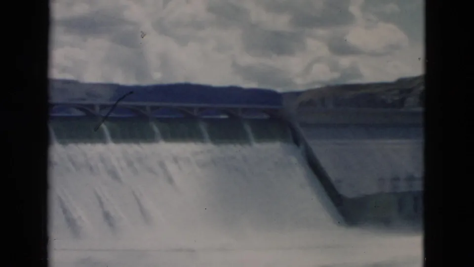 1958 Montana home-movie frame showing spillway water release across a concrete dam, partially obscured by a dark frame edge.