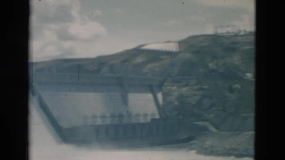 Soft 1958 Montana 8mm frame showing the dam structure, hillside, water below, and a pale building-like form on the slope.