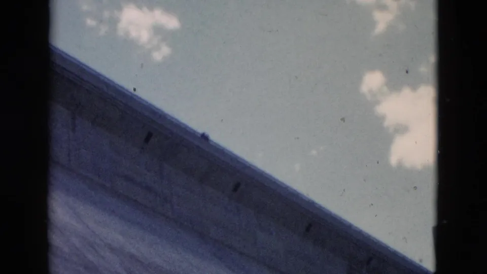 Low-angle 1958 8mm view of a broad concrete dam or bridge surface rising diagonally beneath blue sky and small clouds.