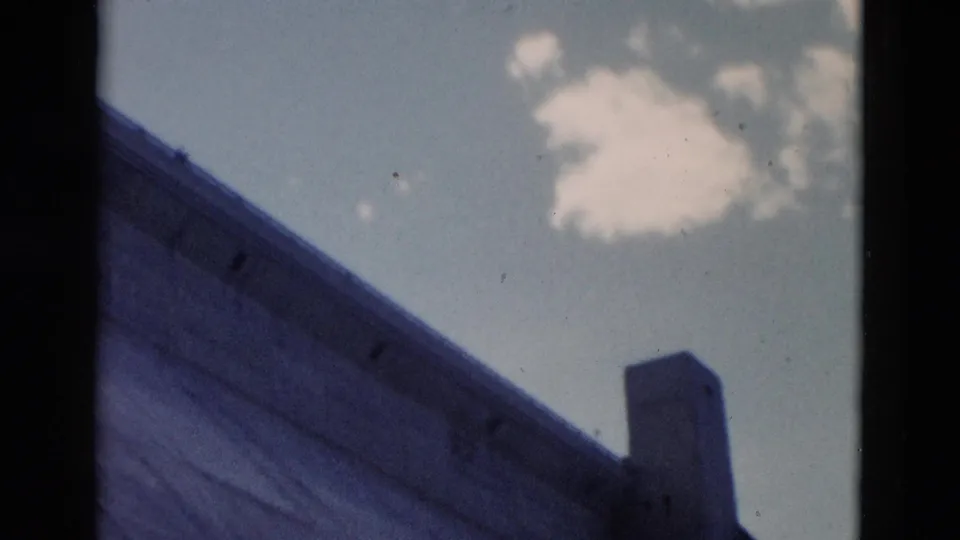 1958 Montana home-movie frame of the dam or bridge structure from below, showing a vertical concrete element against bright sky and clouds.