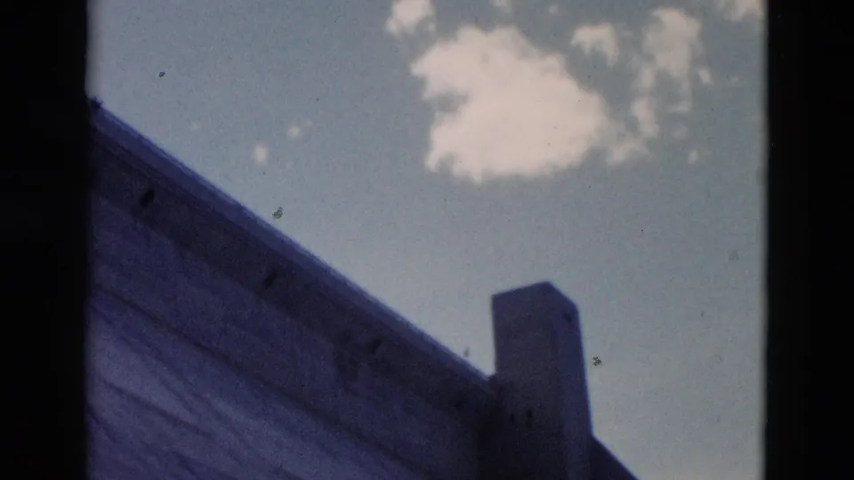 1958 Montana 8mm frame looking upward at a sloped built edge and pillar-like form against pale blue sky with a small white cloud.