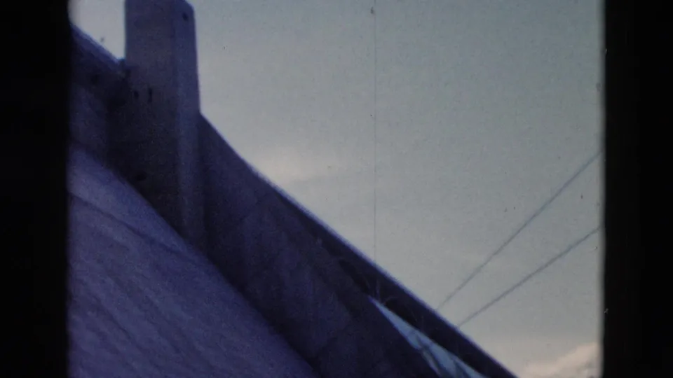 1958 Montana 8mm frame showing a steep roof plane, chimney, pale sky, and faint overhead lines.