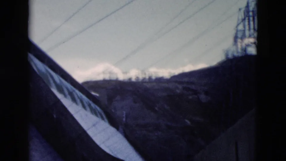 1958 Montana frame showing a roof edge, overhead wires, snowy peaks, hills, and a tower-like structure at right.