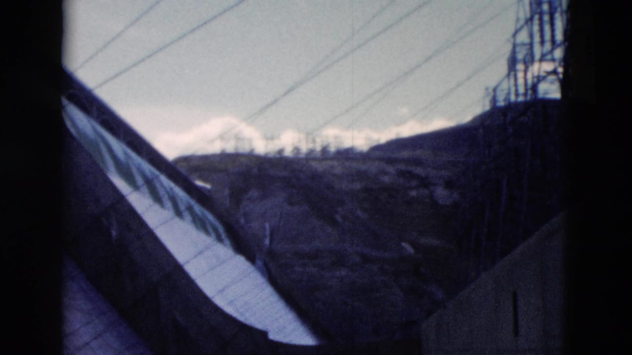 Curved concrete dam wall with power lines and mountain terrain beyond.