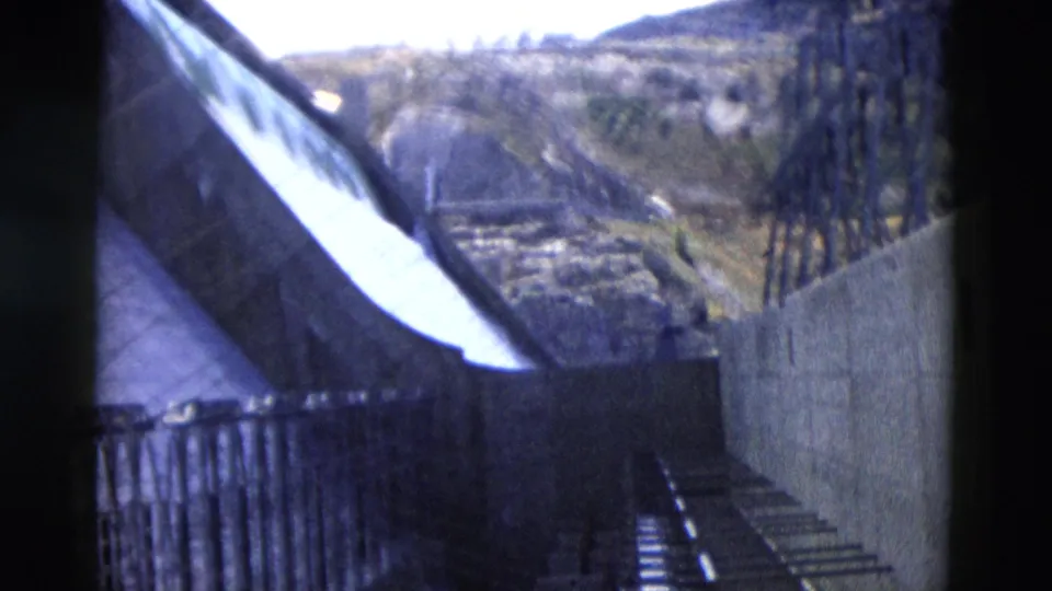 1958 Montana 8mm frame showing the dam wall, sloped spillway edge, railings, concrete supports, and rugged hills beyond.
