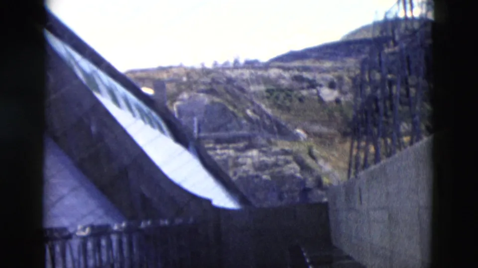 1958 Montana 8mm frame looking past a roofline or barrier toward a sloped hillside and tall utility structure.