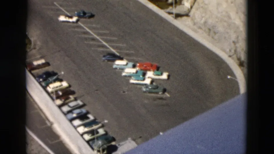 1958 Montana 8mm frame of a parking lot from above, with clustered cars, striped spaces, and hillside terrain at the edge.