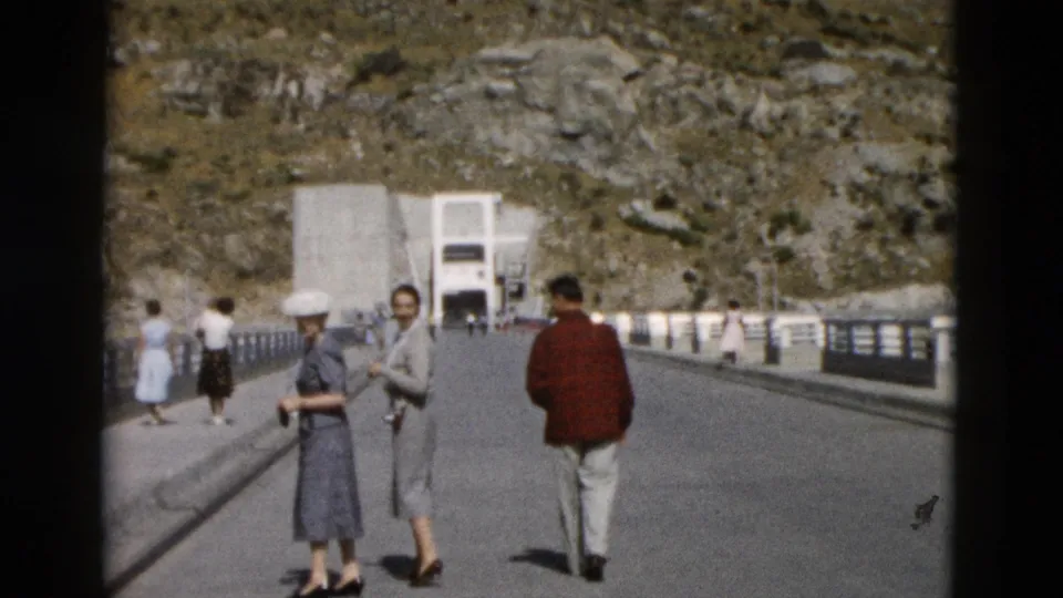 1958 Montana home-movie frame with visitors walking on a bridge, including women in hats and a man in a red jacket.