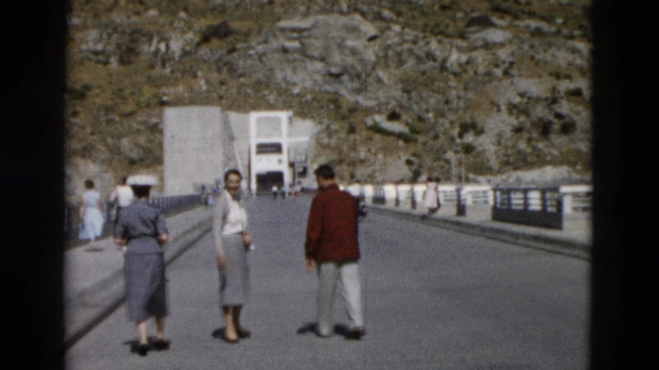 Adults standing and walking on a wide bridge toward a large concrete structure in a rocky mountain setting.