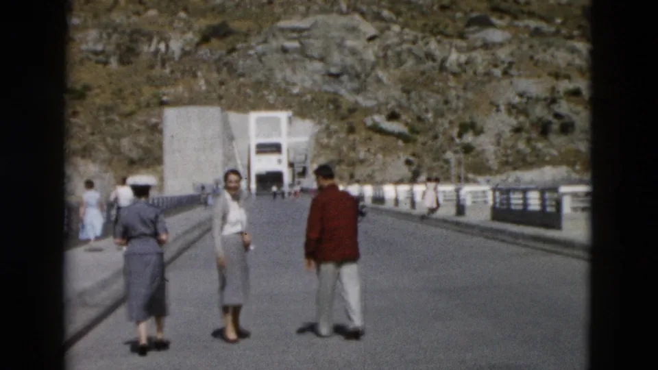1958 Montana home-movie frame showing visitors on a broad bridge leading toward a large concrete structure below rocky slopes.