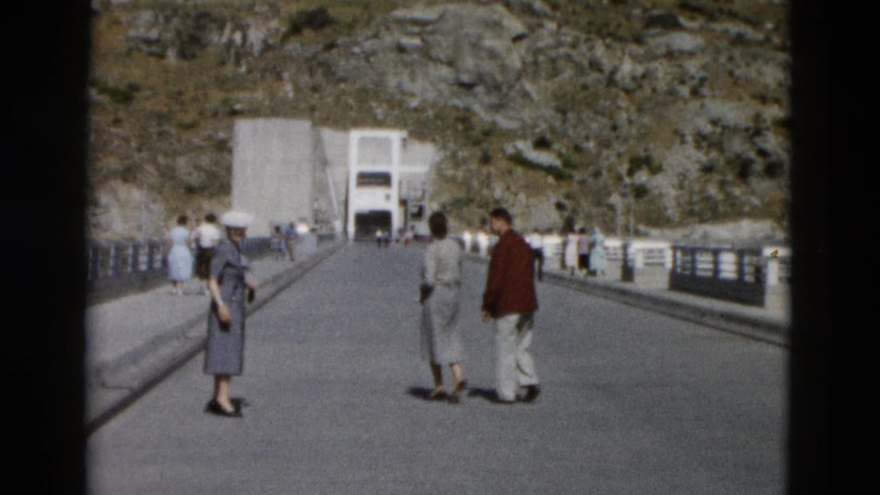 Several adults walk away along a broad bridge with railings toward a concrete structure in the distance.