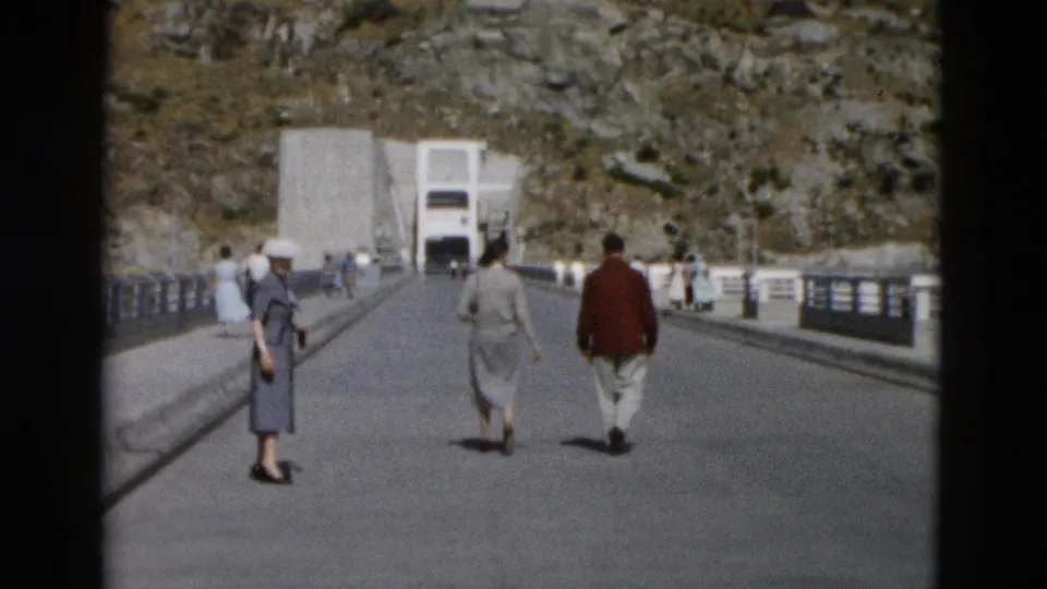 1958 Montana travel footage frame with adults moving away on a bridge toward a concrete structure set against rocky terrain.