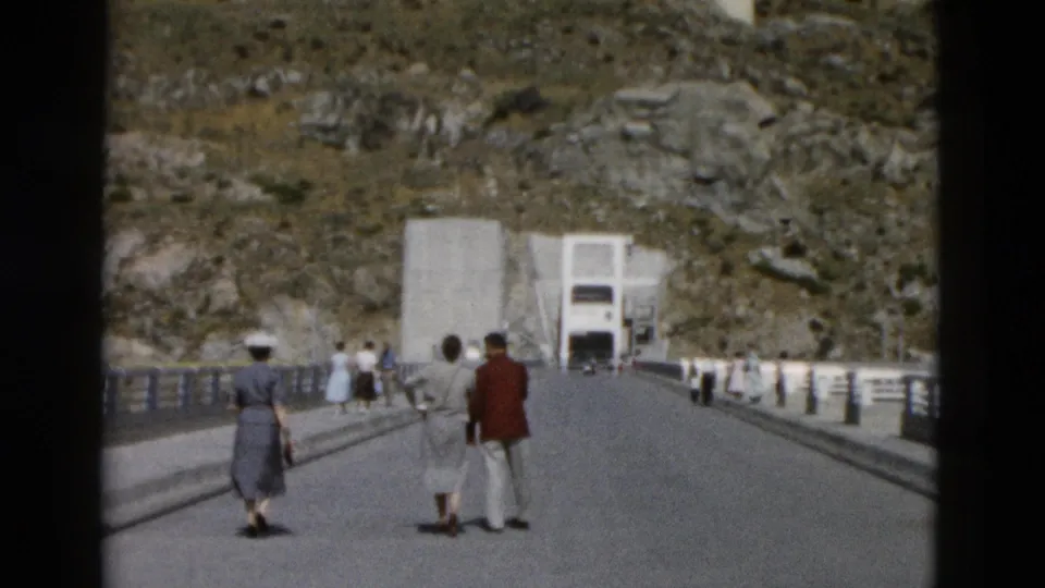 Visitors walk away from the camera on a wide Montana bridge, approaching a concrete dam-like structure in 1958 home-movie footage.