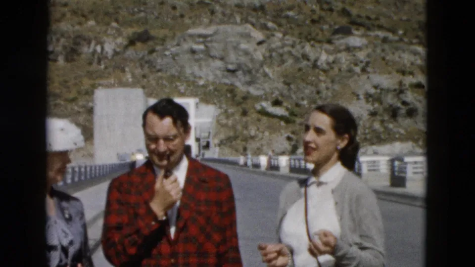 1958 Montana home-movie frame of adults on a sunny mountain road, including a man in a red plaid jacket and women beside him.