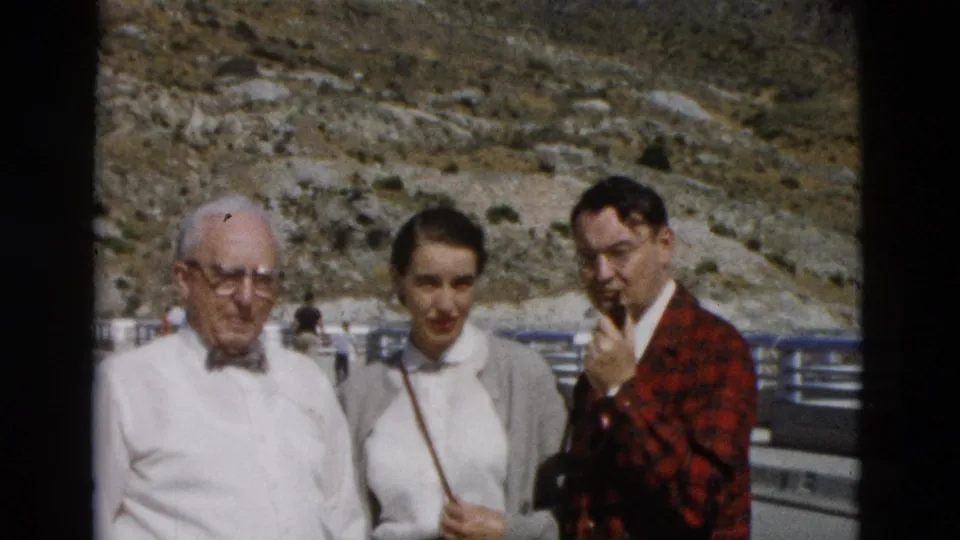 1958 home-movie frame showing an older man, a woman in a grey cardigan, and a man in a red plaid jacket near a mountain road railing.