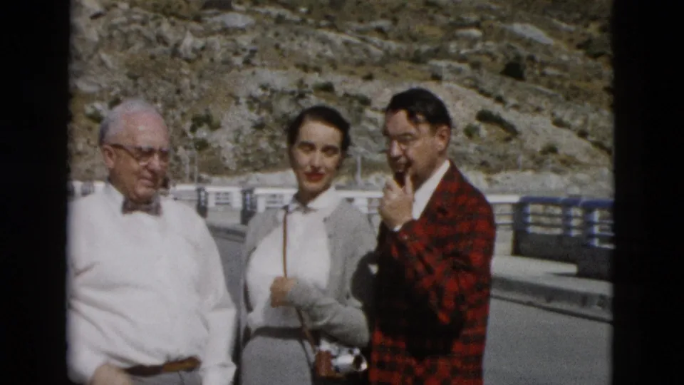 The group faces the 8mm camera beside a railing, while the man in the red plaid jacket raises a hand to his chin.
