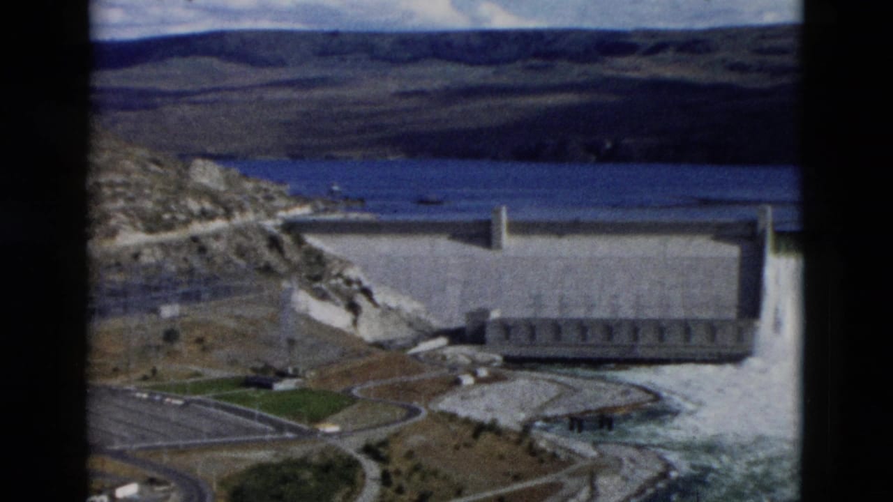 A large concrete dam spans water with a reservoir and hills beyond.