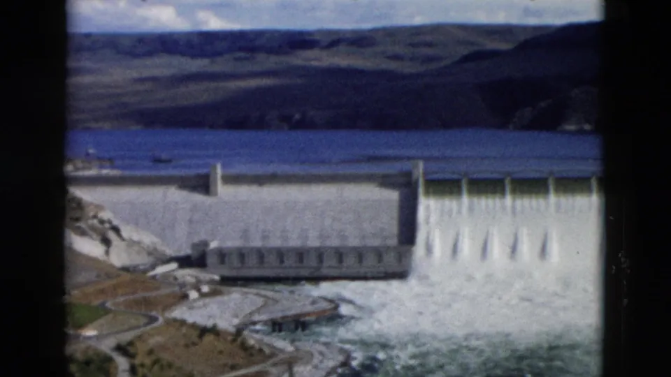 A closer 1958 Montana home-movie view shows heavy spillway flow from a concrete dam, with reservoir water and hills behind.