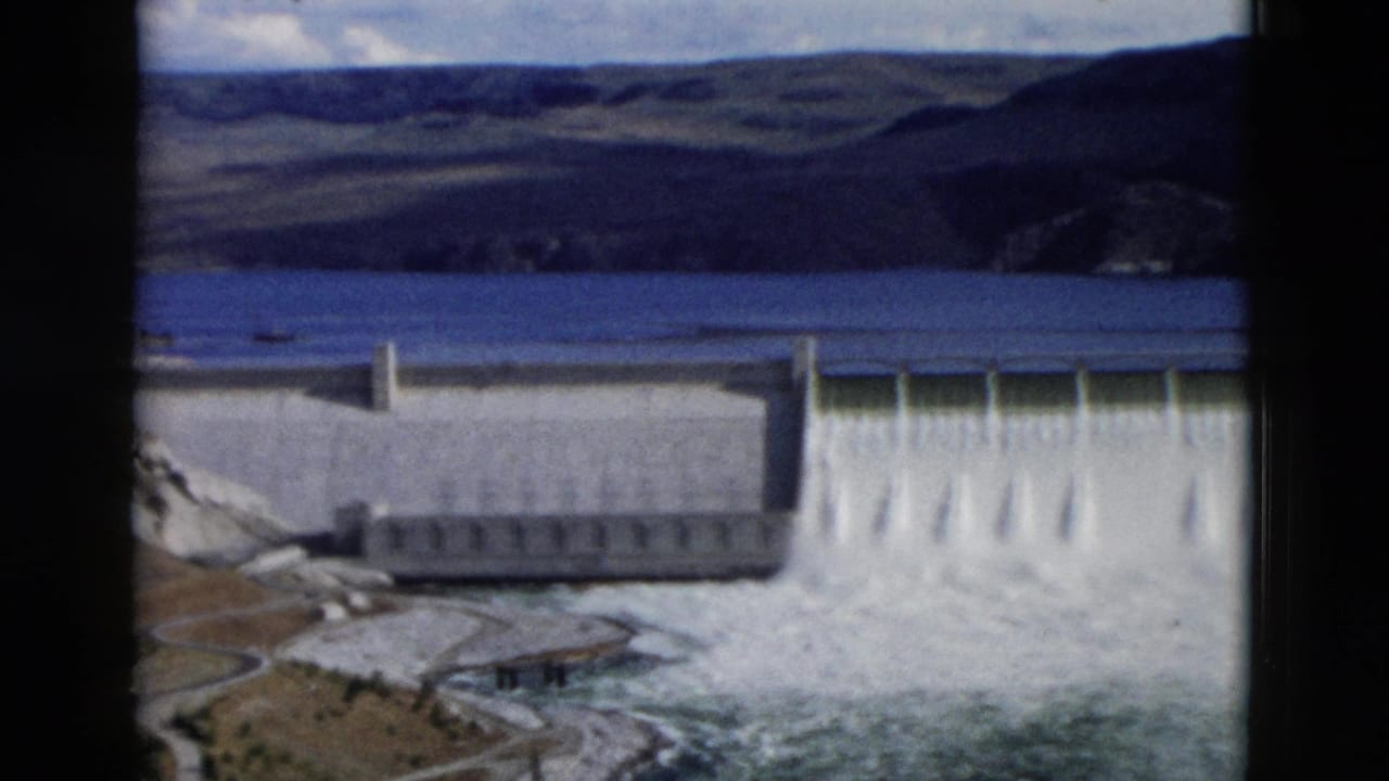 Concrete dam spillway releasing water beneath a reservoir and hills.